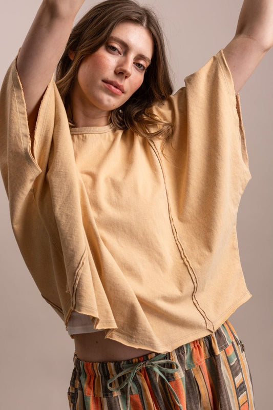 A woman wearing a loose, oversized short-sleeve top in a warm sandy beige color, paired with colorful striped pants, posing with her arms raised against a solid neutral background.