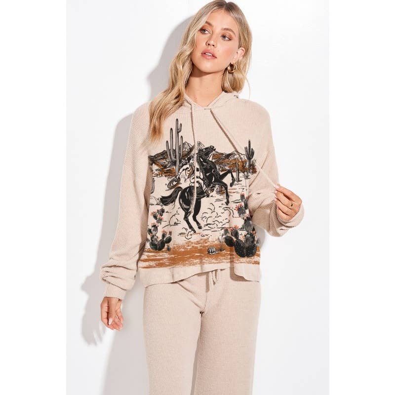 A woman modeling a beige hooded sweatshirt and matching sweatpants set. The hoodie features a large graphic print on the front depicting a cowboy riding a horse with cacti and desert scenery.