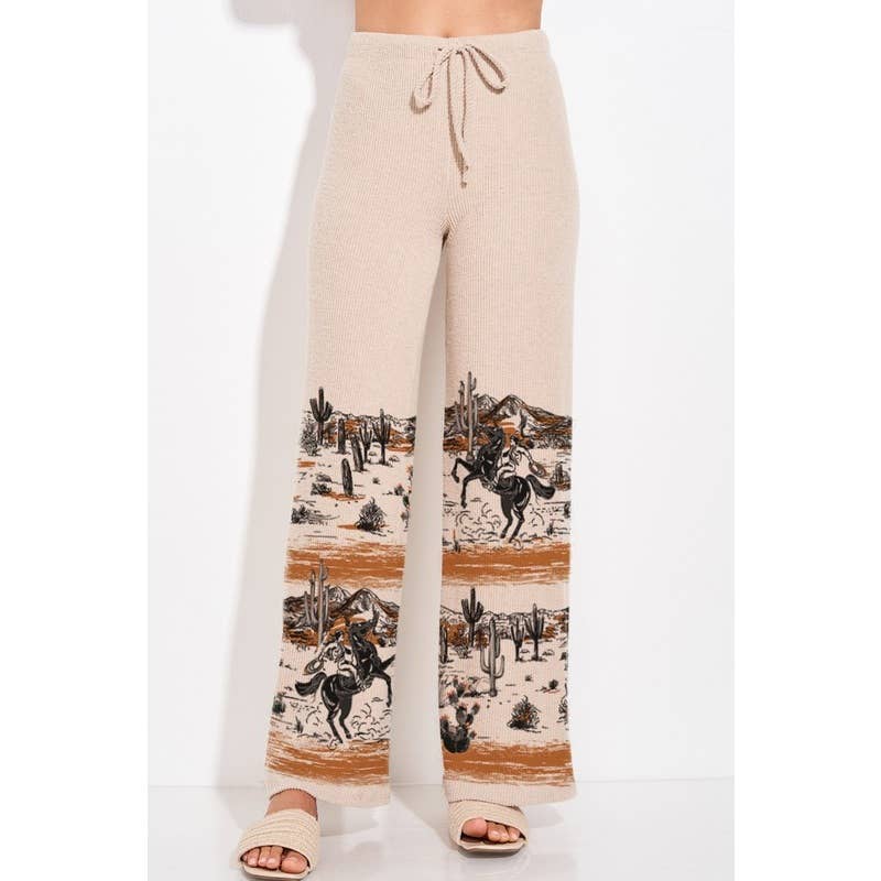 A person wearing beige wide-leg knit pants with a desert cowboy print near the lower legs, shown against a plain white studio background. The image is cropped from just below the chest to the feet, and the person is wearing beige slide sandals.