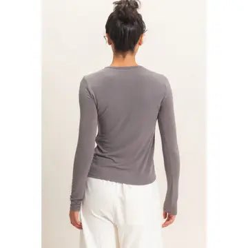 A woman shown from the back wearing a fitted long-sleeve crewneck top in a muted gray color, paired with white pants, against a plain light background.