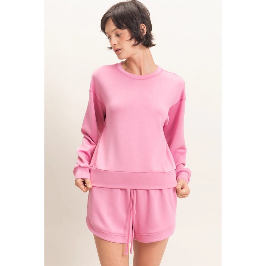 A woman modeling a matching pink lounge set consisting of a long-sleeve sweatshirt and drawstring shorts against a plain light background.
