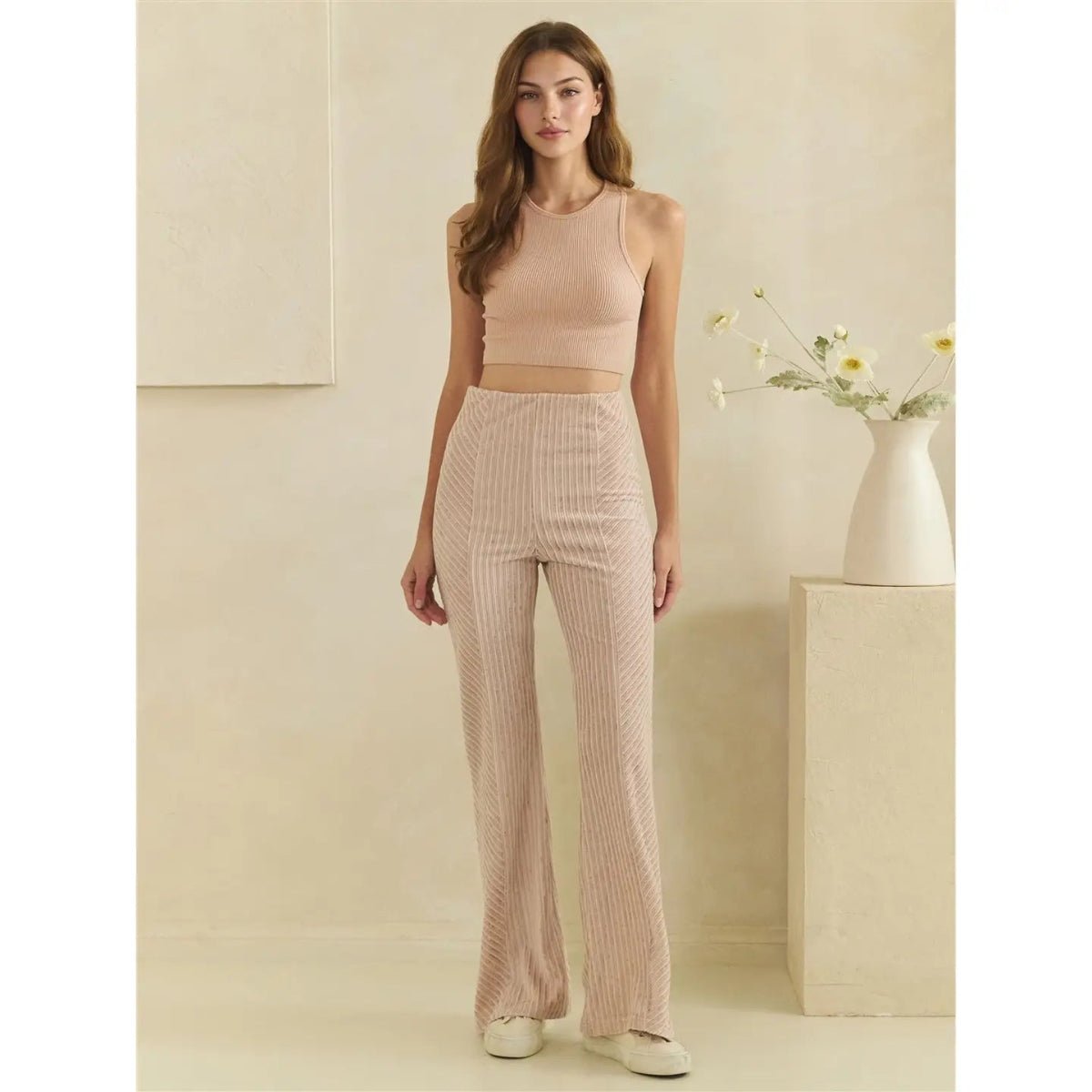 A woman stands in a minimalist studio setting wearing a matching ribbed knit two-piece set consisting of a sleeveless crop top and high-waisted wide-leg pants in a light peach-beige color. She is standing next to a white vase with small yellow flowers on a beige pedestal.