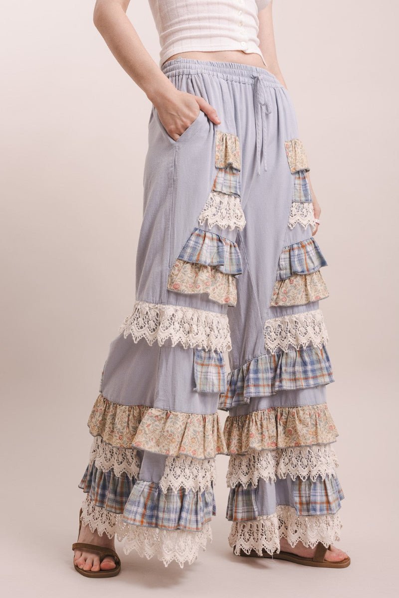 A person wearing loose, wide-leg ruffled pants in a dusty lavender-gray fabric with layered plaid and cream lace ruffle details, paired with a white cropped top and sandals, against a plain beige studio background.