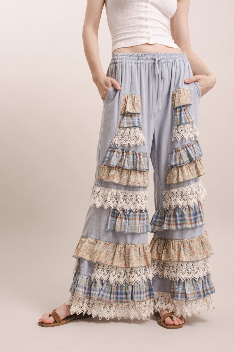 A model wearing light blue wide-leg ruffled pants with layered plaid and lace details, paired with a white ribbed top and brown sandals, against a solid beige studio background.