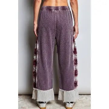 A person wearing loose, wide-leg purple pants with white frayed fringe at the hem and matching fringe running down the sides, shown from the waist down against a plain indoor wall.