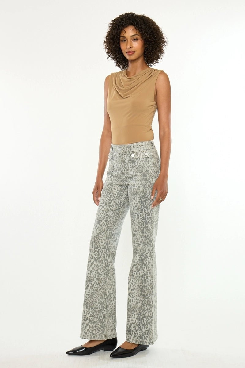 A model stands against a plain white studio background wearing a tan sleeveless cowl-neck top and high-waisted, wide-leg pants with a gray and white abstract pattern. She is wearing black pointed flats.