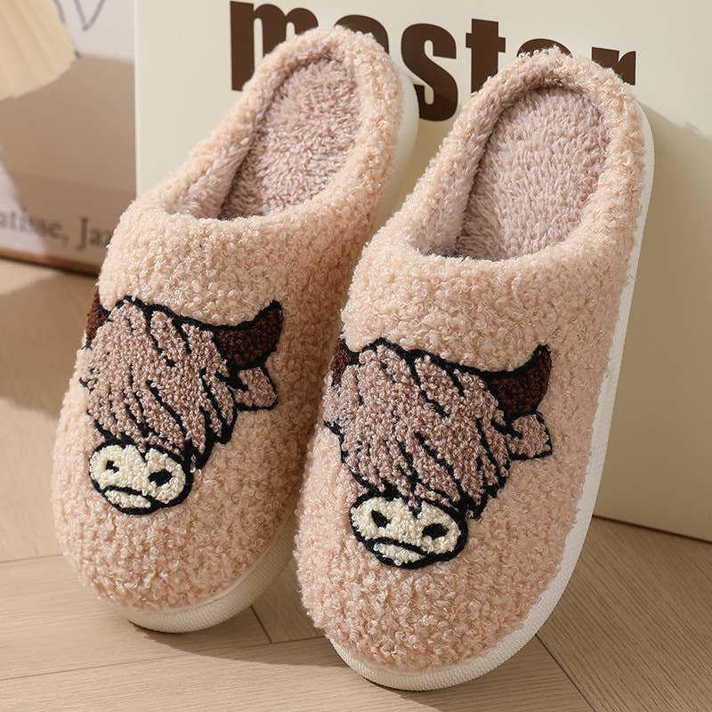 A pair of plush fleece slip-on house slippers in a light peach-pink color, featuring embroidered cartoon cow faces on the front and white soles, shown on a wooden floor.