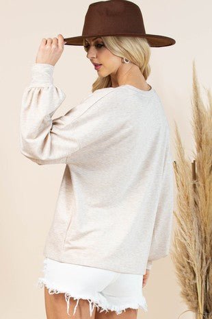 A woman wearing a light beige oversized long-sleeve sweatshirt, white frayed denim shorts, and a brown wide-brim hat, shown from the back against a warm beige background with dried grass decor.