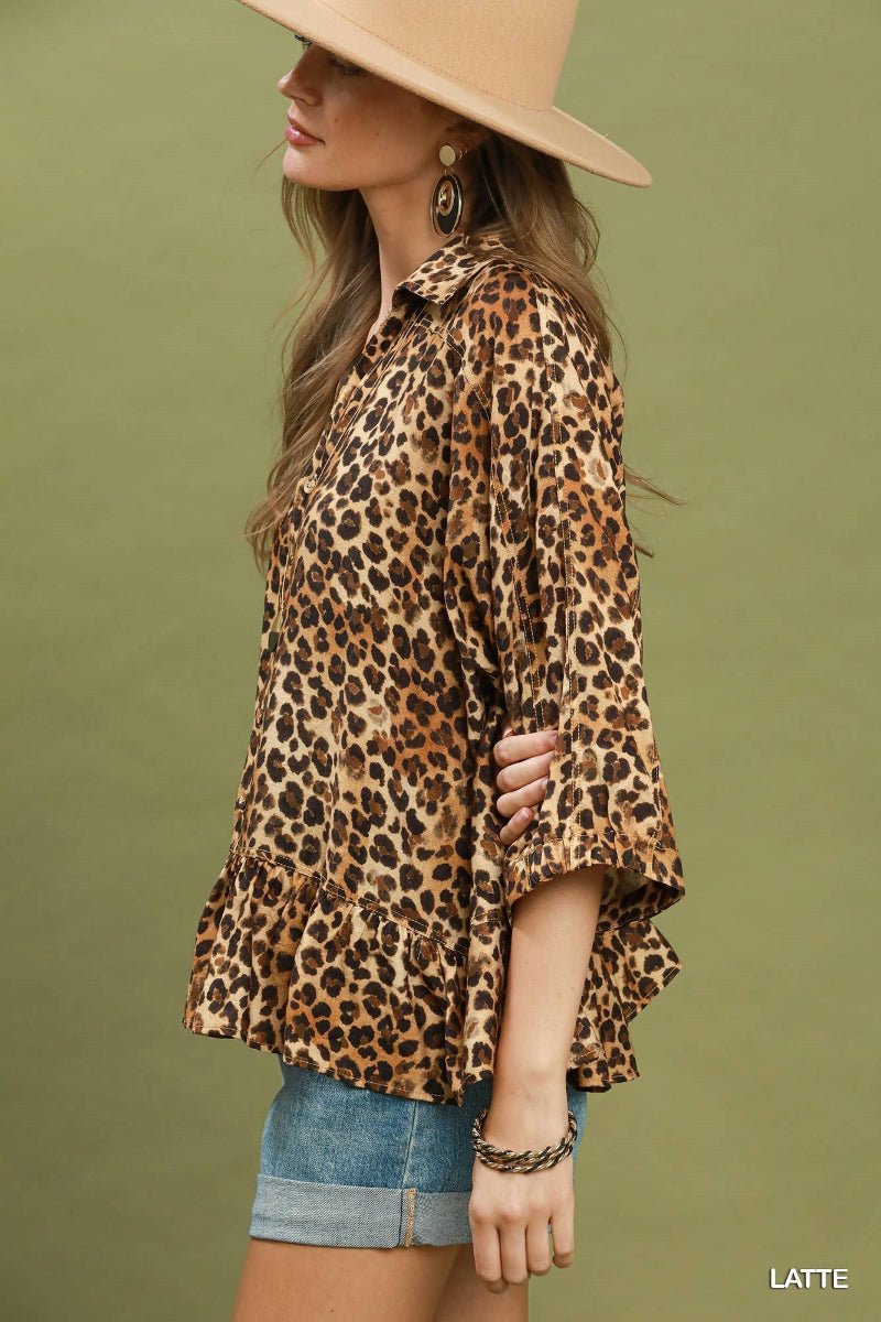 A woman wearing a leopard-print blouse with three-quarter ruffled sleeves and a ruffled hem, paired with denim shorts and a wide-brimmed beige hat, set against a solid olive green background.