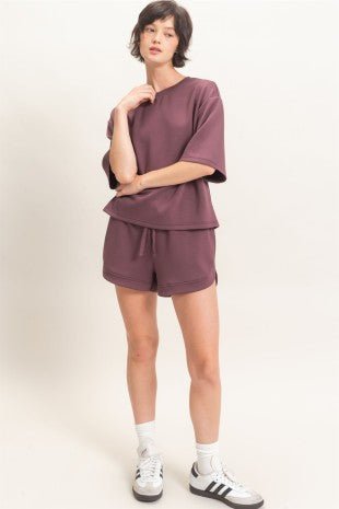 A model wearing a matching mauve-purple lounge set consisting of a short-sleeve crewneck top and drawstring shorts, styled with white socks and white sneakers with black stripes, against a plain off-white studio background.