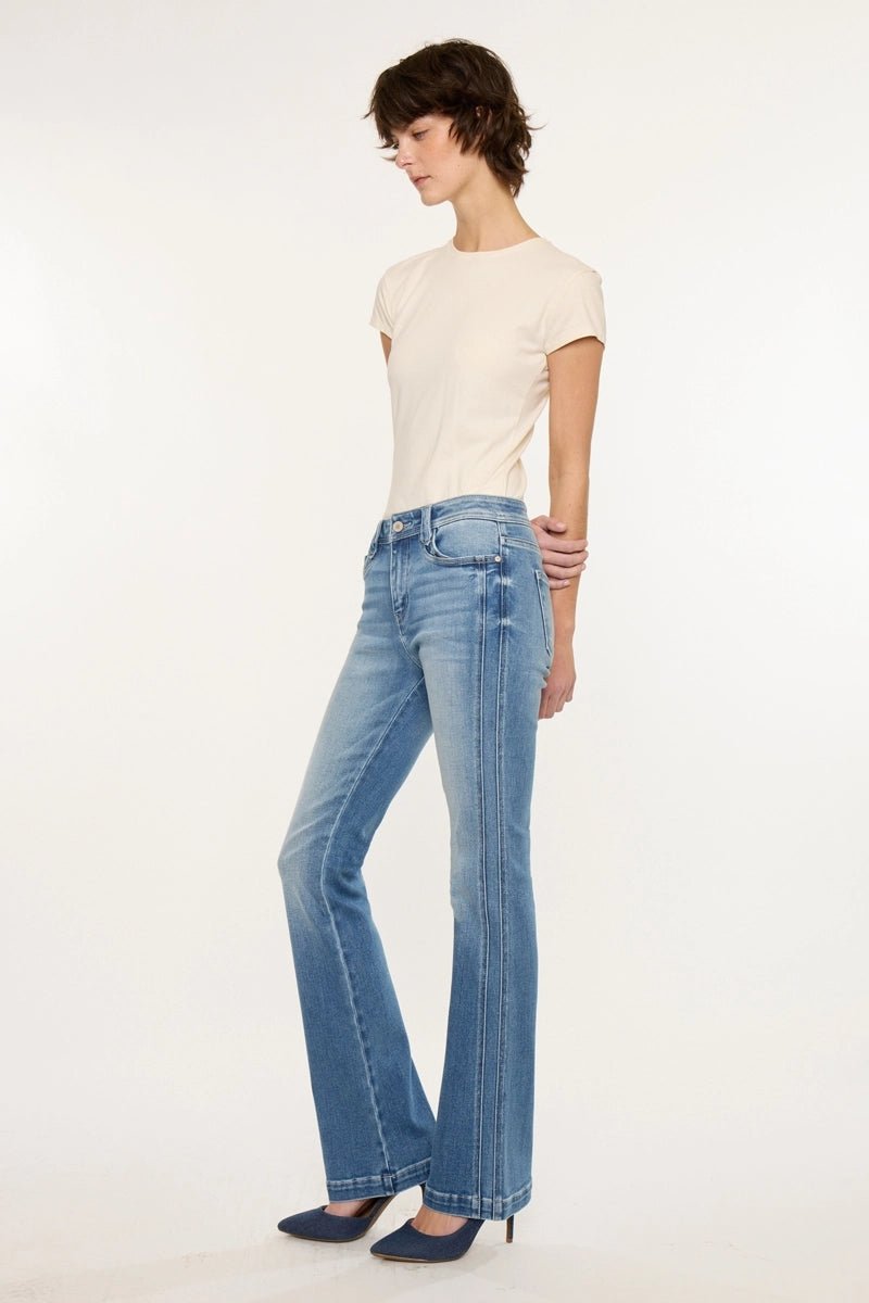 A model wearing light-wash blue flare jeans with distinctive vertical contrast stitching down the outer legs, styled with a cream short-sleeve t-shirt and dark blue pointed-toe heels against a plain white studio background.