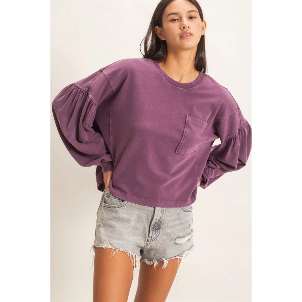 A woman modeling a purple cropped long-sleeve sweatshirt with balloon sleeves and a chest pocket, paired with light-wash distressed denim shorts against a plain light background.
