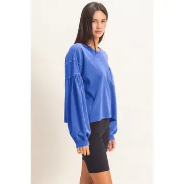 A woman modeling a bright blue long-sleeve blouse with gathered, balloon-style sleeves, paired with black shorts, against a plain off-white studio background.