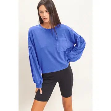 A woman modeling a blue long-sleeve crewneck sweatshirt with a chest pocket, paired with black bike shorts, against a plain light background.