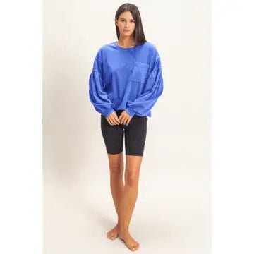 A woman modeling an oversized royal blue long-sleeve top with balloon sleeves and a chest pocket, paired with black bike shorts, against a plain off-white studio background.