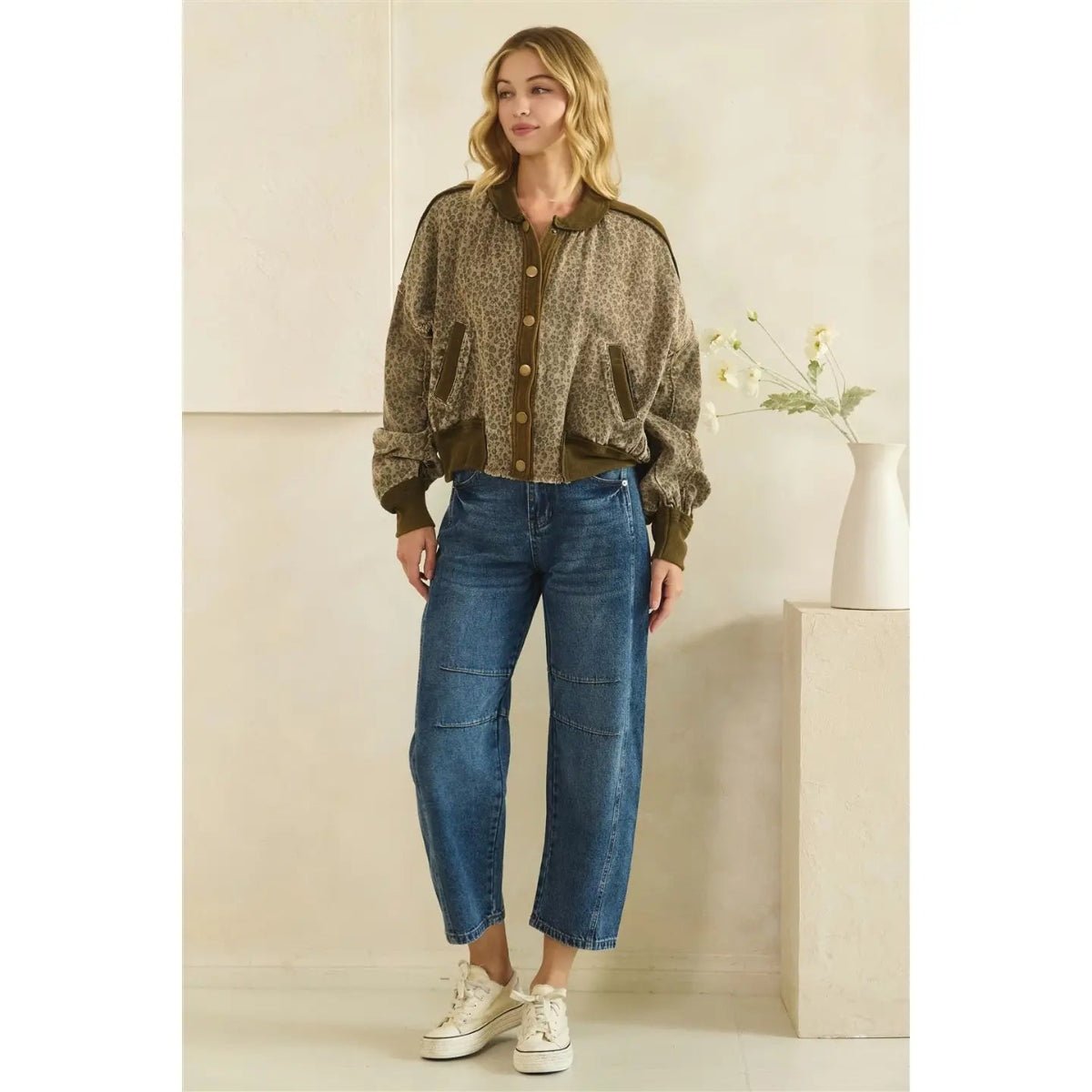 A woman stands indoors wearing a textured olive-brown button-front jacket with wide lapels and front pockets, blue cropped jeans, and white sneakers. The background is a warm beige interior with a small vase of flowers on a pedestal.