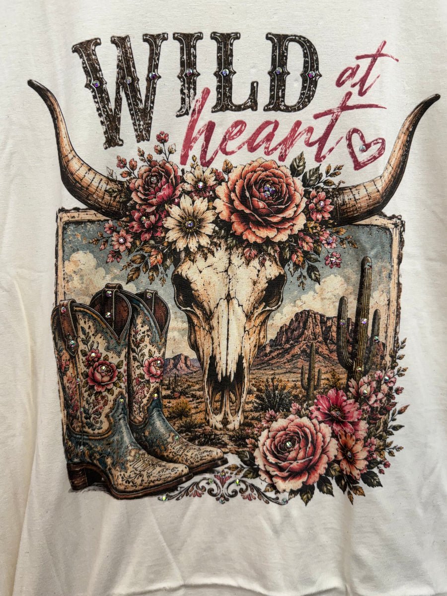 A close-up of a white graphic t-shirt featuring a western design with the text 'WILD at heart', a steer skull with a floral crown, cowboy boots, and a desert landscape.