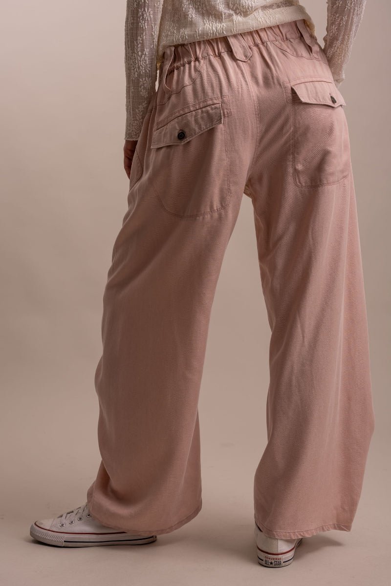 A person shown from the back wearing loose, wide-leg dusty pink pants with flap pockets, paired with a cream knit top and white sneakers, against a plain beige studio background.