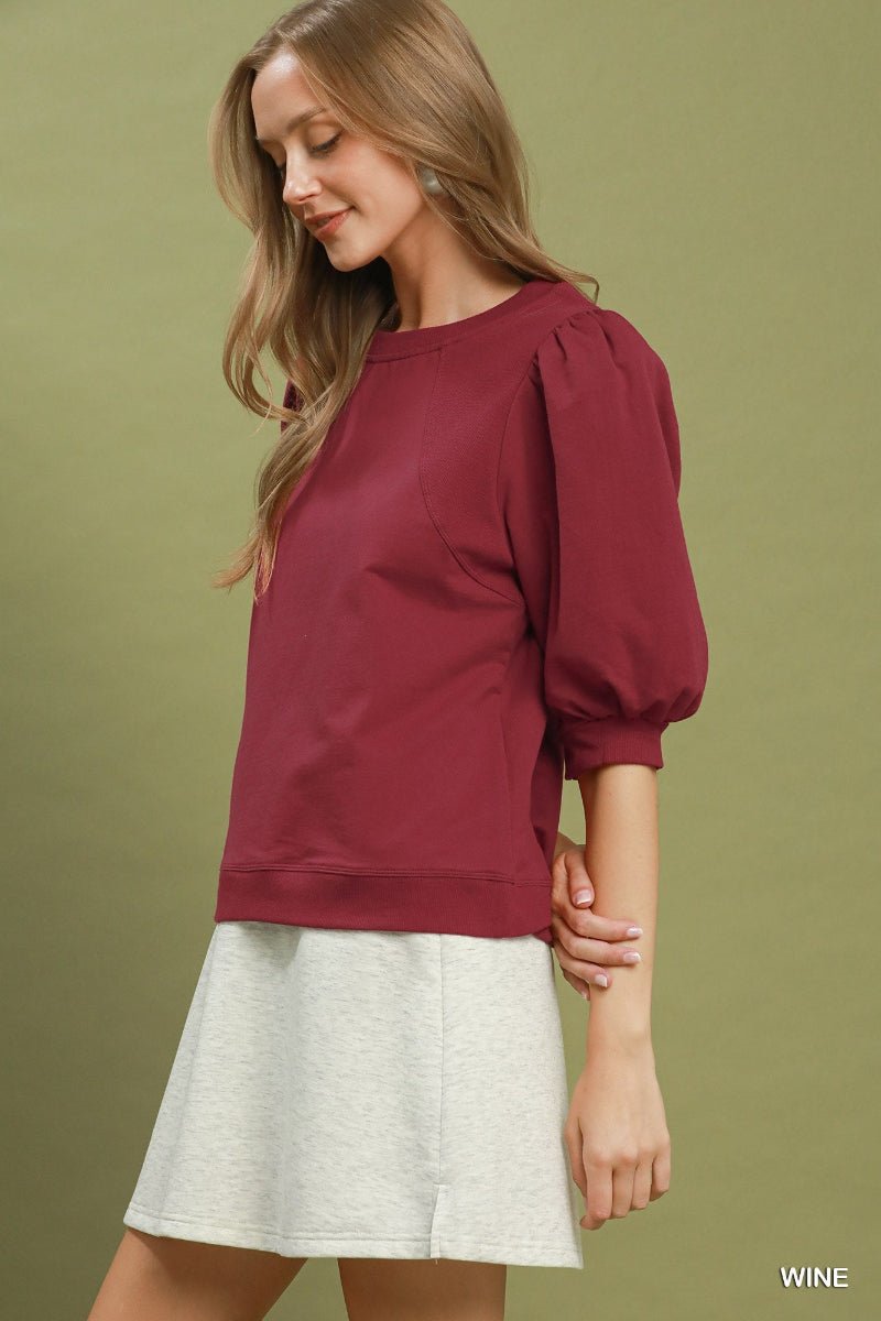 Woman wearing a burgundy top with puff sleeves and a white skirt on a green background