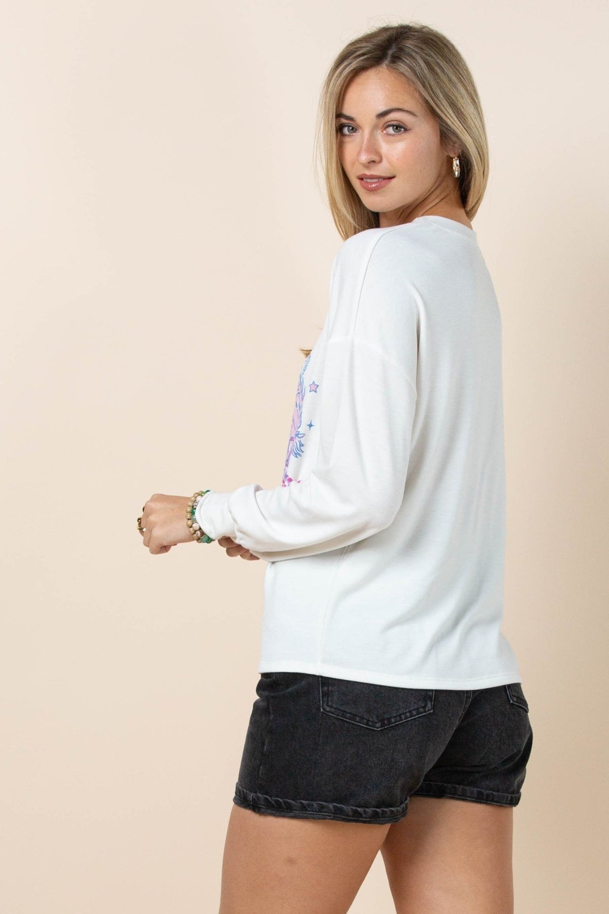 A woman modeling a white long-sleeve crewneck sweatshirt with a colorful graphic on the back, paired with black denim shorts, against a solid beige background.
