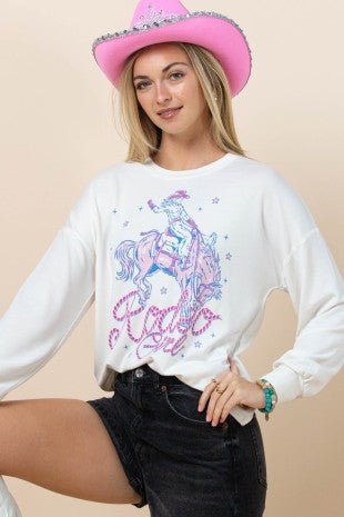 A woman wearing a white long-sleeve crewneck sweatshirt with a pink and blue western-themed graphic of a cowboy on a horse, styled with a pink cowboy hat and dark denim shorts against a solid beige background.