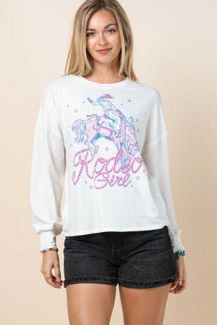 A woman modeling a white long-sleeve crewneck sweatshirt with a colorful graphic of a cowboy on a horse and the text 'Radio Girl' in pink and blue, paired with black denim shorts against a solid beige background.