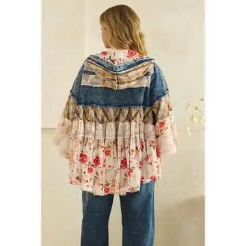 A woman viewed from behind wearing a bohemian-style tiered patchwork cape jacket with floral and denim patterns, paired with blue jeans, against a neutral beige wall.