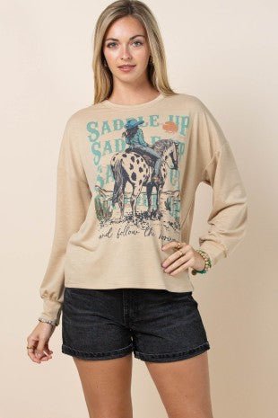 A woman modeling a beige long-sleeve crewneck sweatshirt with a western-themed graphic print of a cowboy on a horse and the text 'SADDLE UP', paired with dark denim shorts against a solid beige background.