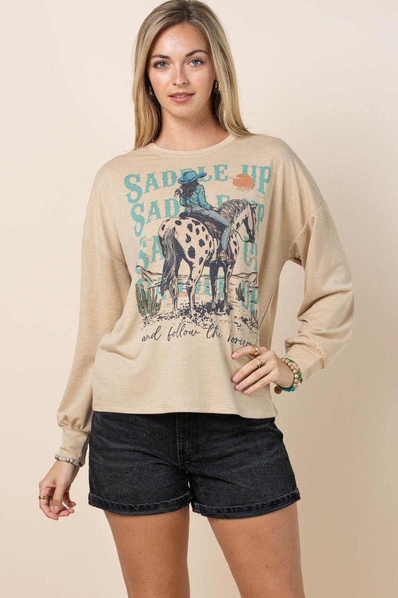 A woman modeling a beige long-sleeve crewneck sweatshirt with a western-themed graphic print of a rider on a horse and the text 'SADLE UP', paired with dark denim shorts against a solid beige background.