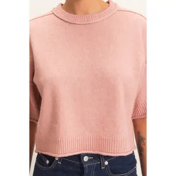 A person wearing a cropped pink knit sweater with a crew neck and short sleeves, paired with dark blue jeans.