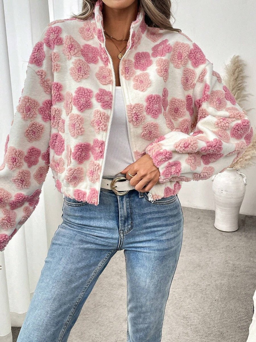A woman wearing a fuzzy white zip-up jacket with pink pom-pom style floral patterns, paired with light blue jeans and a white top, standing in a bright indoor setting.