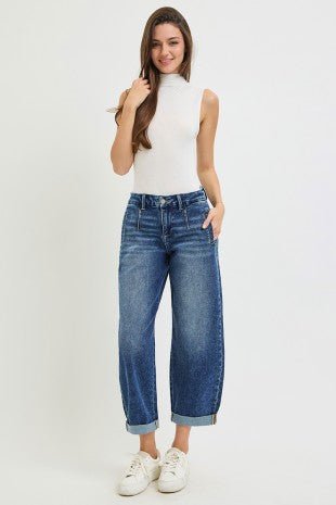 A woman modeling a pair of high-waisted, wide-leg cropped blue jeans with rolled cuffs, paired with a white sleeveless mock-neck top and white sneakers, against a plain white studio background.
