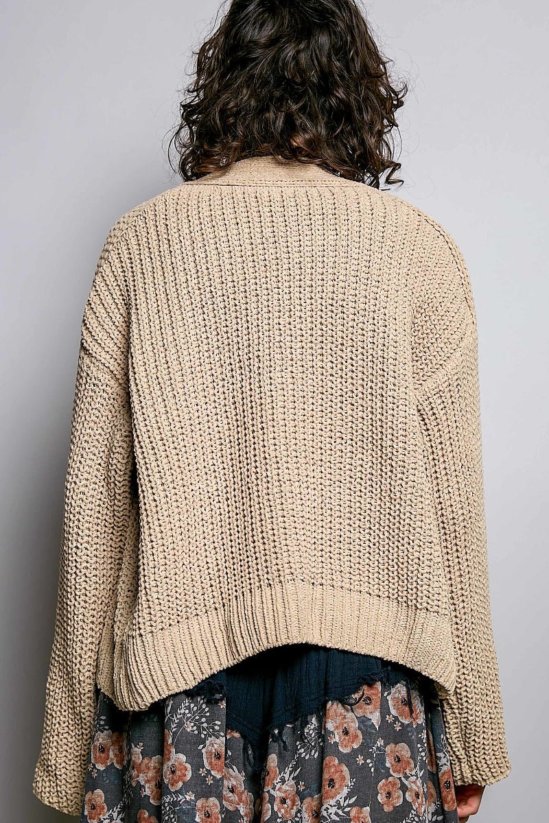 A person shown from the back wearing an oversized beige chunky knit cardigan over a dark floral skirt, against a plain light gray background.
