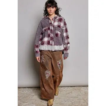 A model wearing a distressed, tie-dye style jacket in gray, white, and dark red, paired with brown wide-leg pants featuring embroidered heart motifs, standing against a neutral studio backdrop.