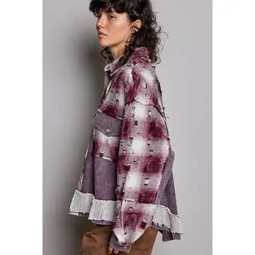 A woman wearing a distressed, oversized plaid shirt jacket in a purple, gray, and white tie-dye pattern, paired with brown pants, shown against a plain gray studio background.