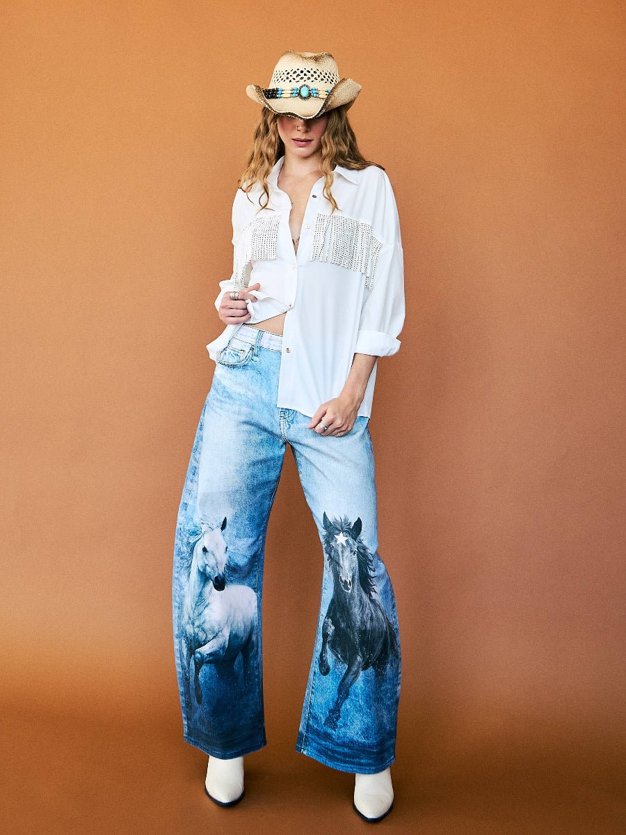 A model wearing a white fringed button-down shirt, wide-leg blue jeans with printed horses, a straw cowboy hat, and white boots, standing against a solid brown studio background.