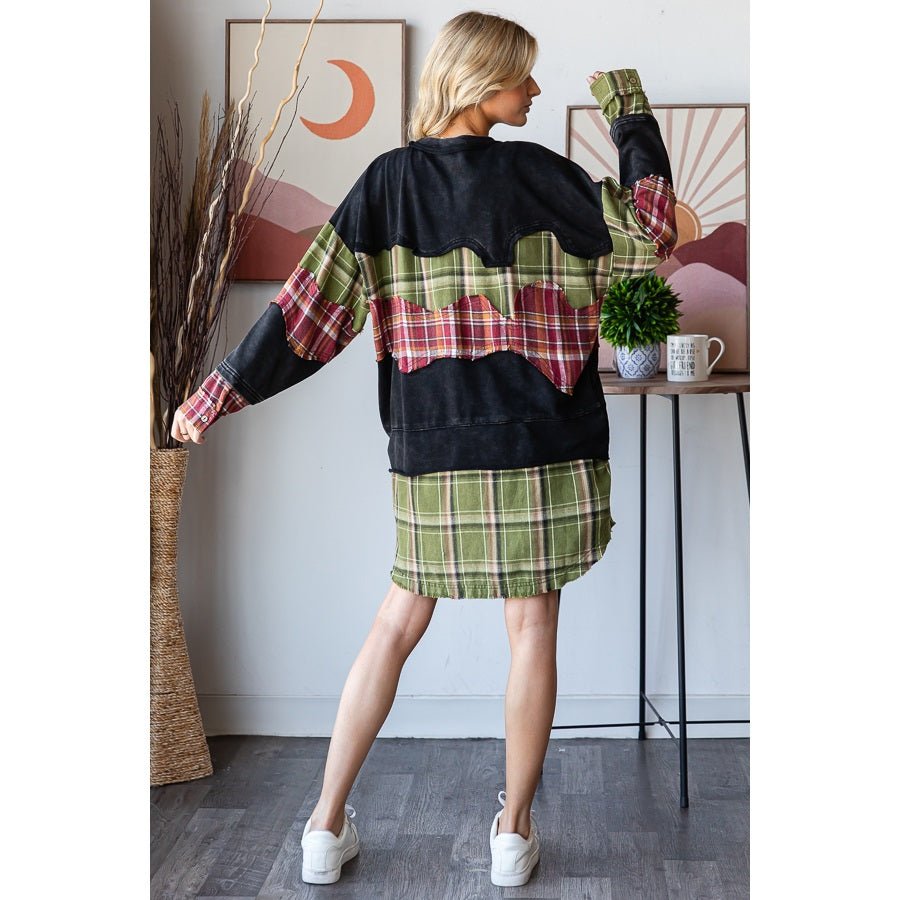 A woman modeling a black oversized sweatshirt with red and green plaid patterned sleeves and hem, paired with a matching green plaid mini skirt, in a styled indoor setting.