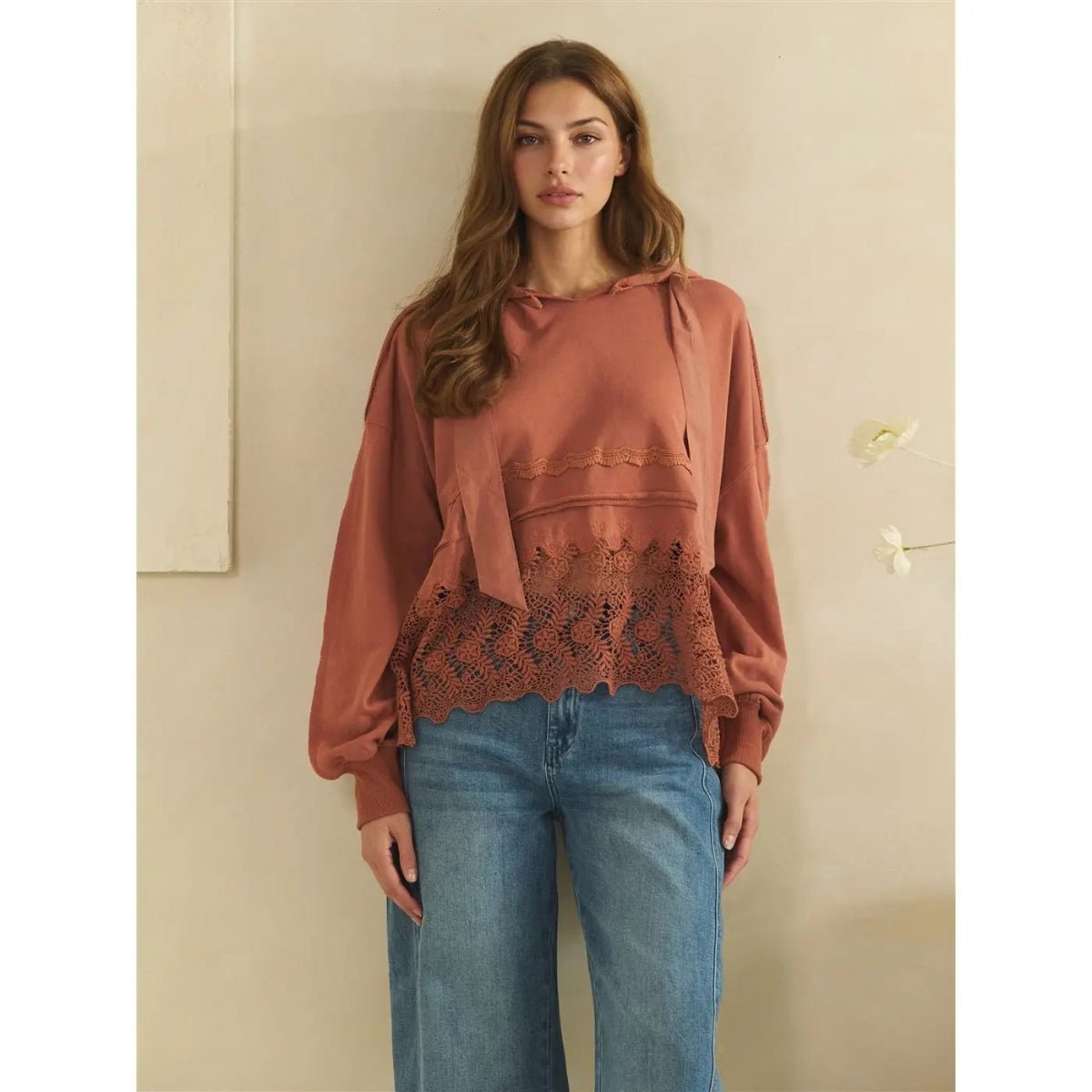 A woman modeling a terracotta-colored long-sleeve blouse with a lace hem and tie detail, paired with blue jeans, standing against a neutral beige wall.