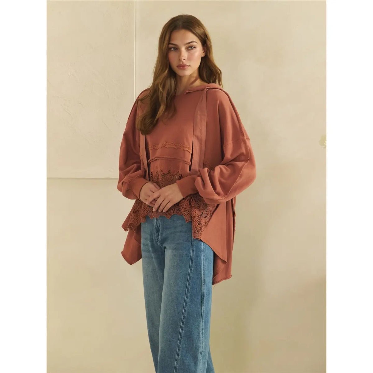 A woman modeling a loose-fitting terracotta-colored blouse with long balloon sleeves and a scalloped lace hem, paired with blue jeans, against a neutral beige wall.