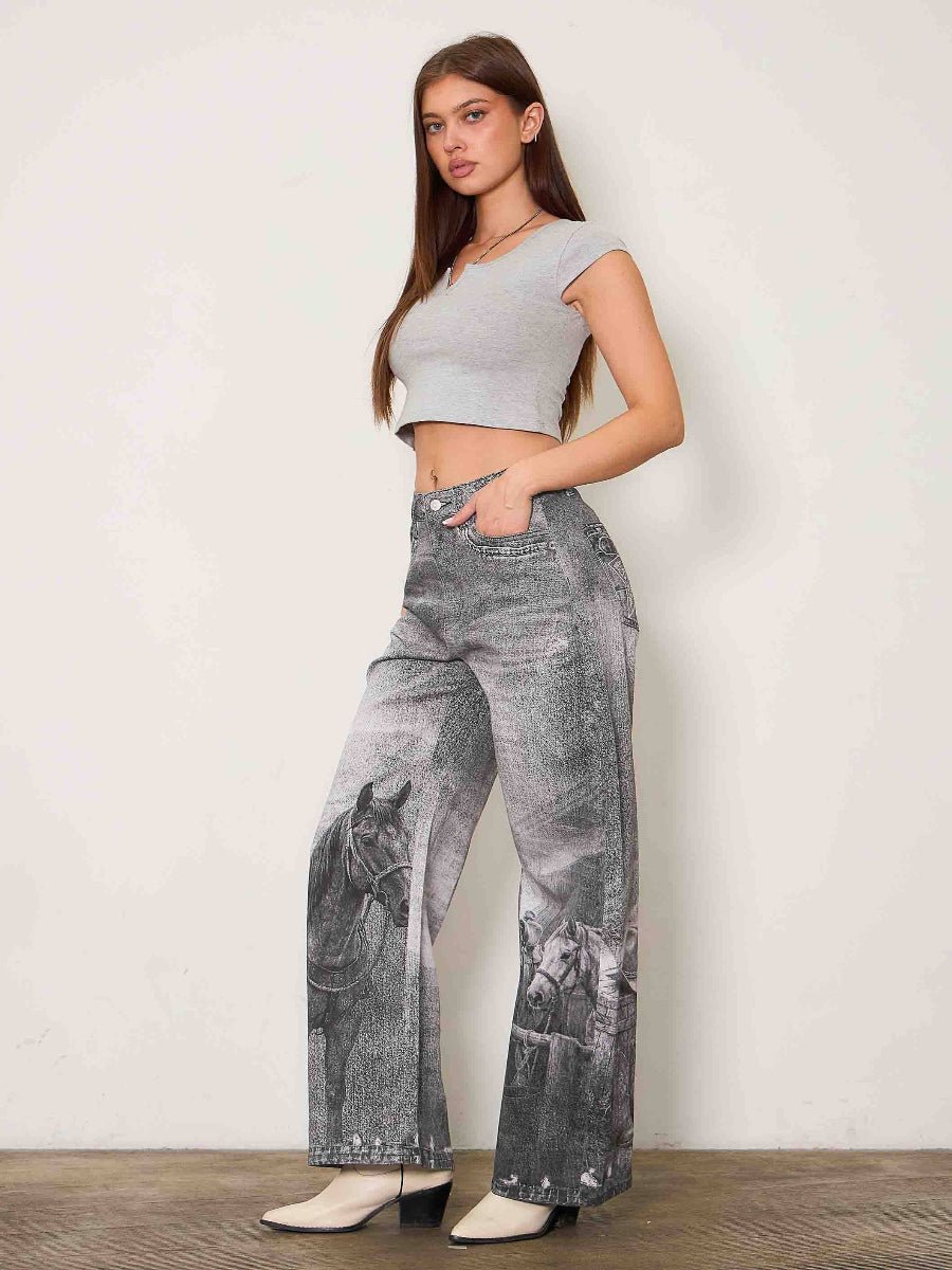 A woman modeling high-waisted, wide-leg gray jeans with a black and white horse print on the lower legs, paired with a light gray crop top and white heeled boots, standing against a plain off-white wall.
