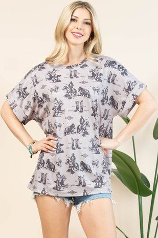 A woman modeling a light gray short-sleeve t-shirt with a black and gray French terry cloth cowboy and rider pattern, paired with denim shorts against a beige background with a green plant.