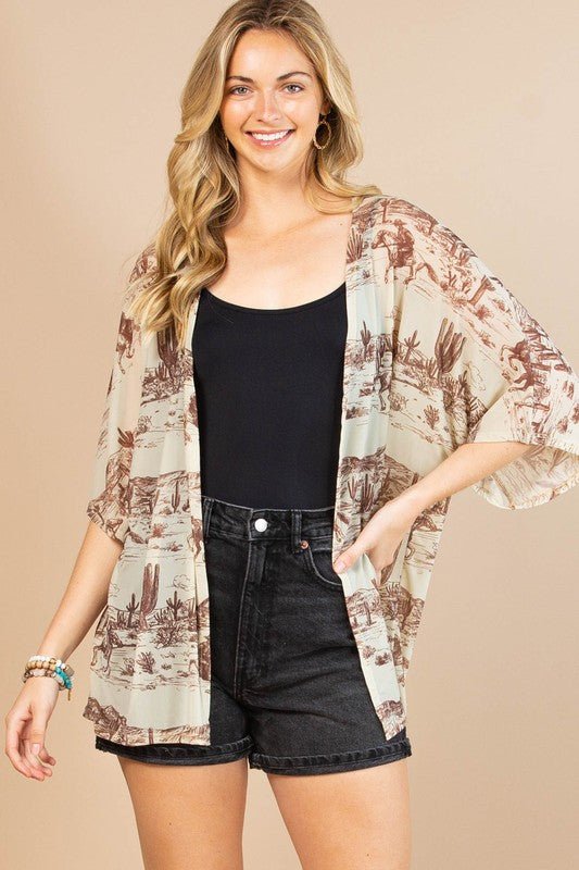 A woman modeling a lightweight open-front kimono-style cardigan with a brown and beige landscape print over a black tank top and dark denim shorts, against a solid beige studio background.