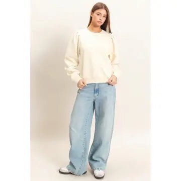 A woman modeling a cream-colored long-sleeve top with puffed shoulders and light-wash wide-leg jeans, standing against a plain off-white studio background.