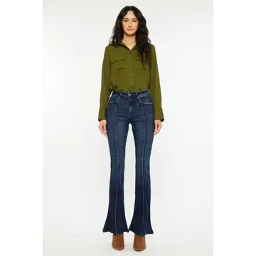 A woman modeling a long-sleeve olive green button-up shirt and dark blue high-waisted flare jeans with brown boots, standing against a plain white studio background.