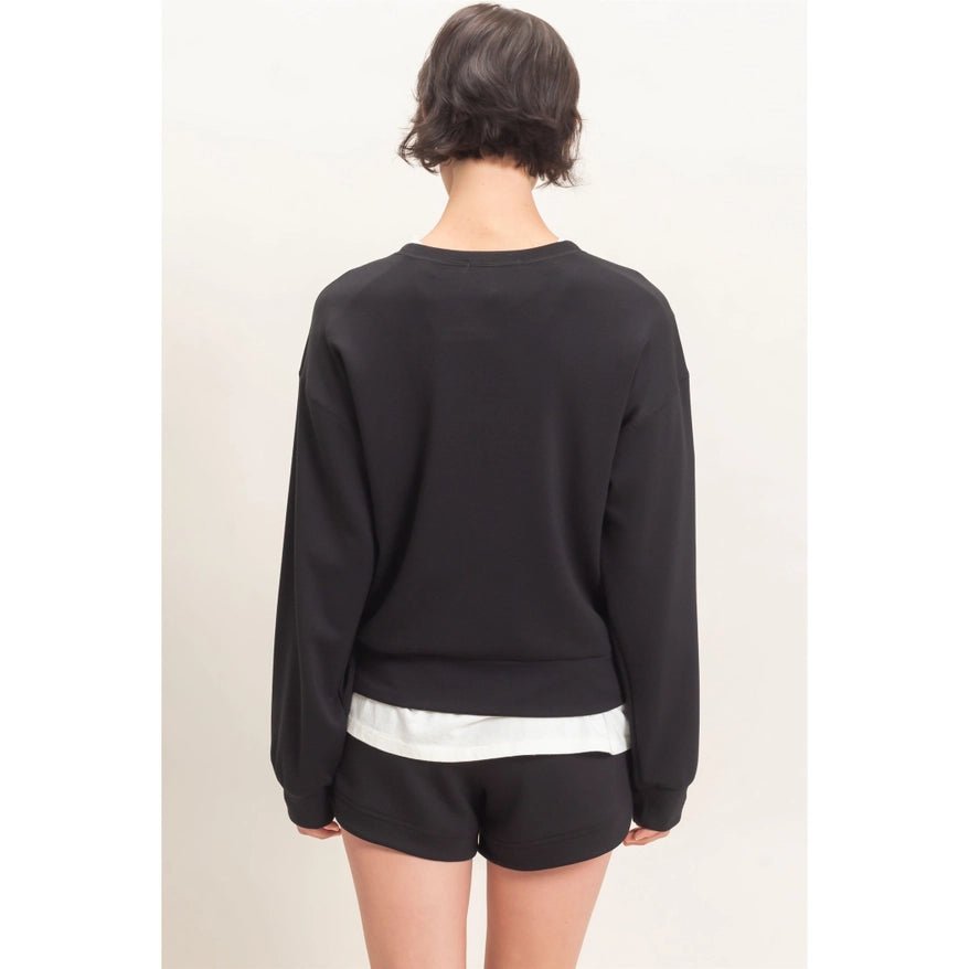 A model shown from the back wearing a black long-sleeve crewneck sweatshirt and black shorts with a white hem detail, against a plain light background.