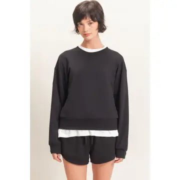 A woman modeling a black crewneck sweatshirt with a white layered hem detail, paired with black shorts, against a plain off-white background.