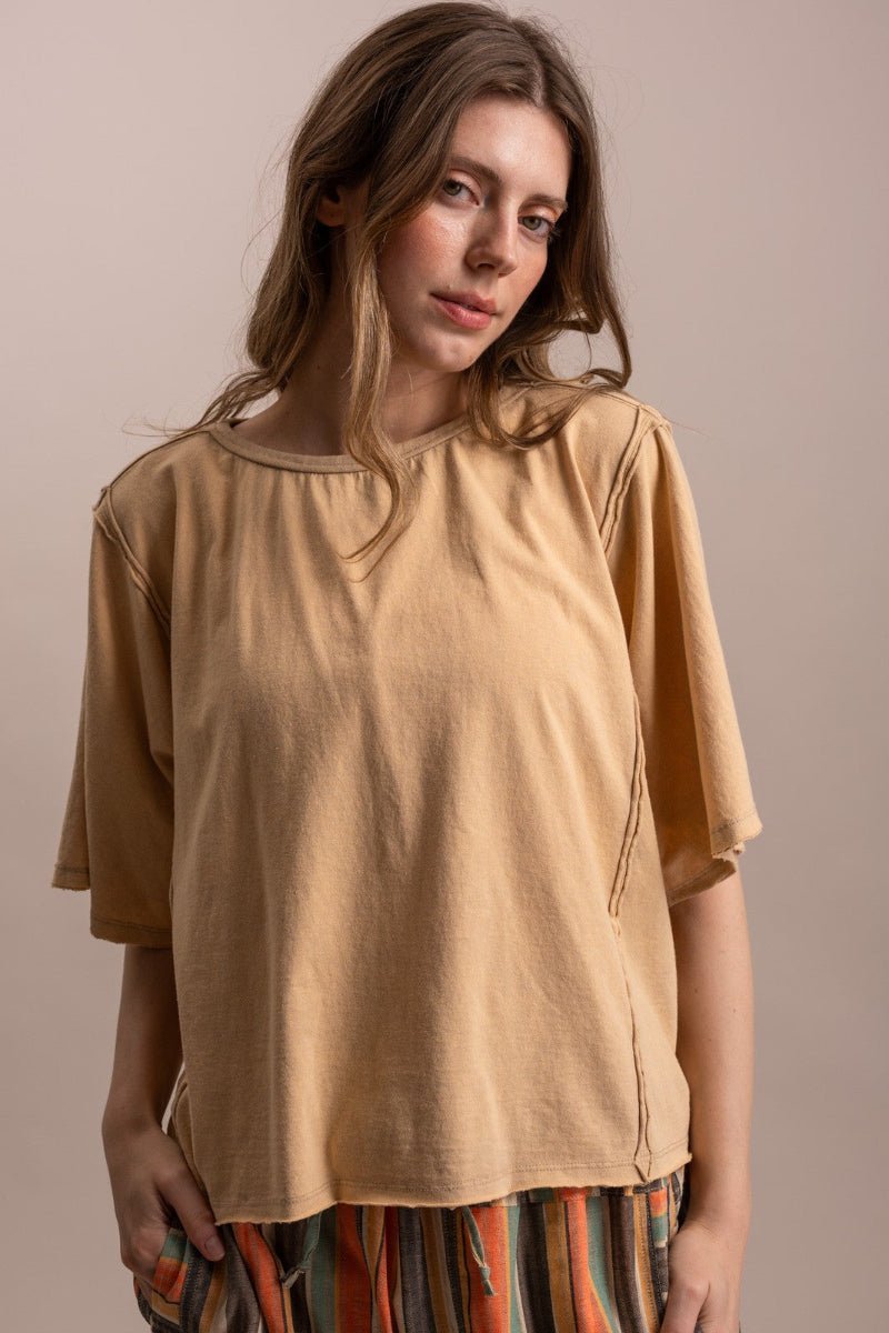 A woman wearing a loose, short-sleeved tan top with side slits and a round neckline, paired with striped pants, against a solid beige background.