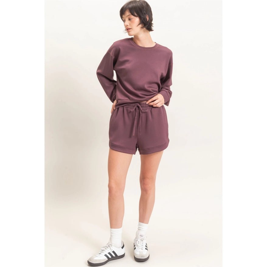 Person wearing a matching maroon sweatshirt and shorts set with white sneakers on a plain background