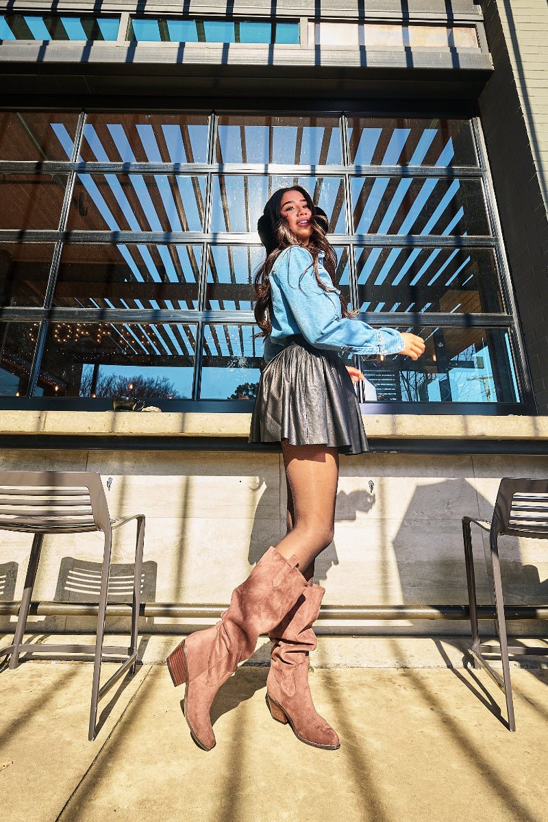 A woman posing outdoors in front of a modern glass building, wearing a light blue long-sleeve top, a dark metallic pleated mini skirt, and dusty rose pink slouchy knee-high boots.