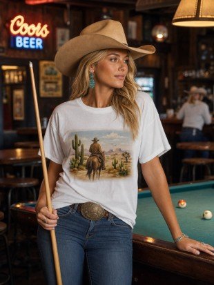 A woman in a cowboy hat and jeans stands in a bar setting, holding a pool cue and wearing a white graphic t-shirt with a western desert scene print. A Coors Beer neon sign is visible in the background.
